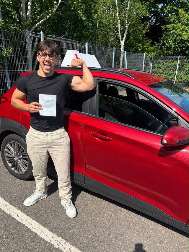 Learner 1 holding their driving test pass certificate beside a red training car