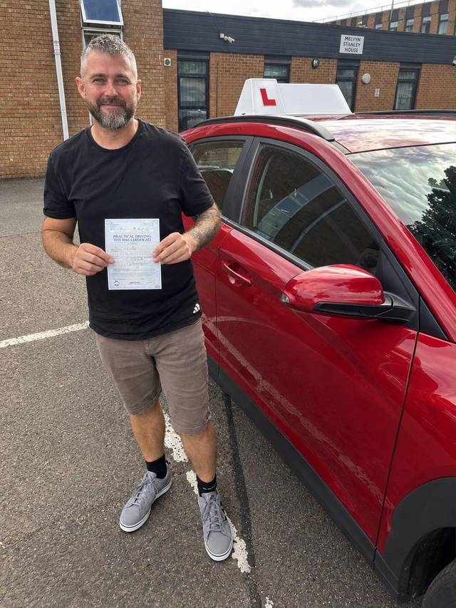 Learner 3 holding their driving test pass certificate beside a red training car